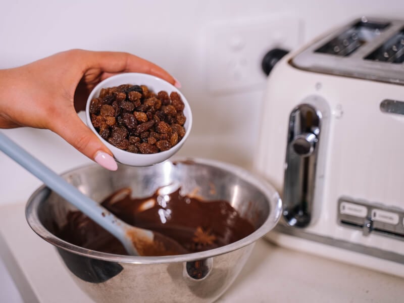 A hand pouring raisins into a bowl of melted chocolate.