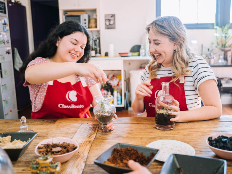 Two women laughing as one sprinkles small pebbles into an up-cycled glass bottle terrarium