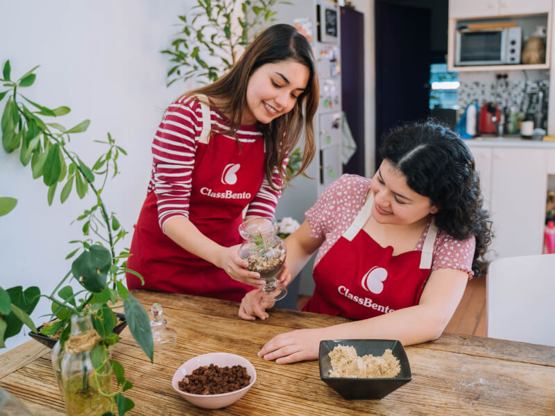 Two women in red aprons admire a finished DIY upcycled glass terrarium containing soil, pebbles and a small green plant