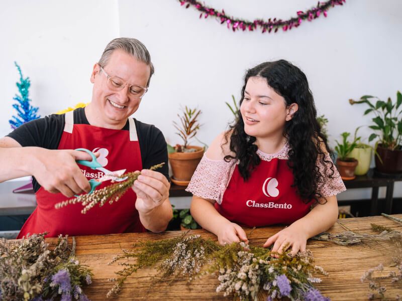 A man trimming pink dried flowers with scissors whilst a woman watches and picks out green and white dried flowers