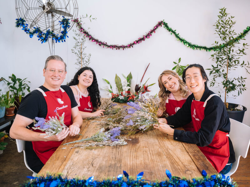 Four people in aprons sitting at a wooden table holding bunches of dried purple and white flowers