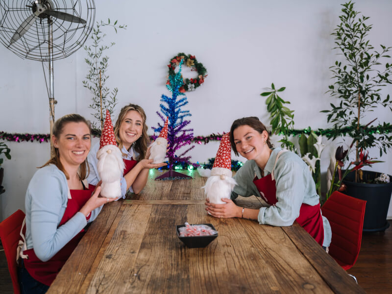 Three women smiling with their DIY Christmas gnome decorations which are wearing red Santa hats
