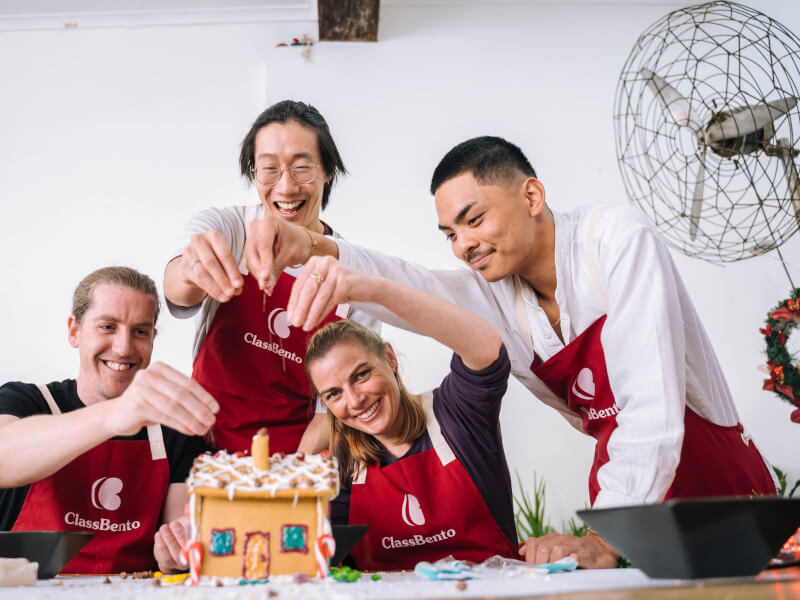 Four people in aprons sprinkle chocolate nibs from a height onto a gingerbread house