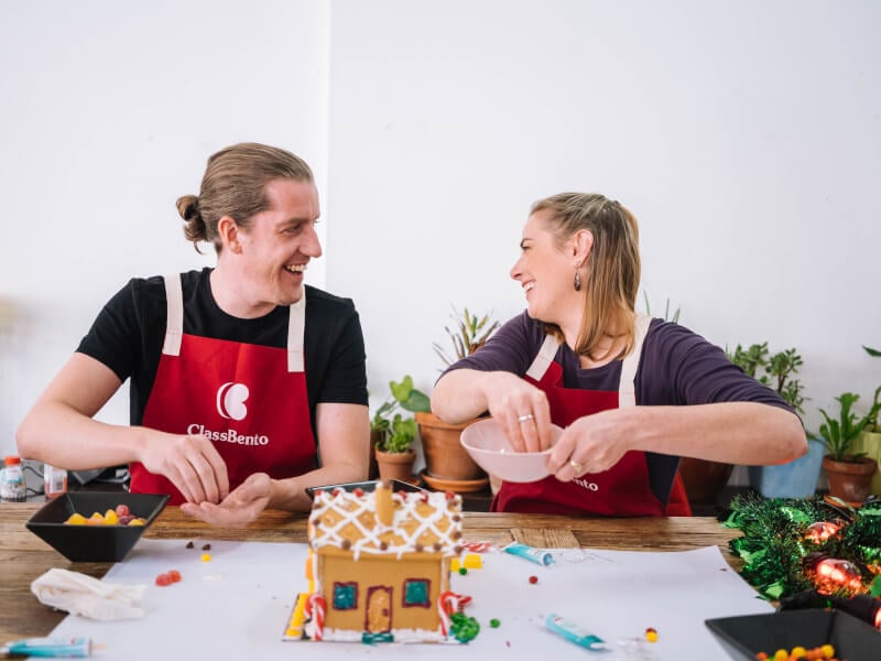 A man and woman, with a gingerbread house in front of them, picking jelly sweets out of dishes