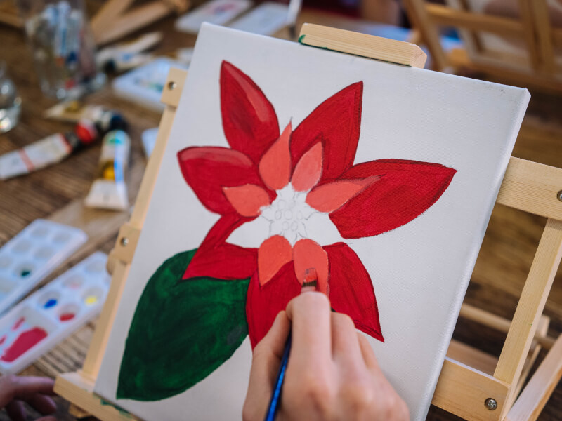 Close up of a hand holding a blue paintbrush to paint a red Christmas flower on a canvas supported by a wooden easel