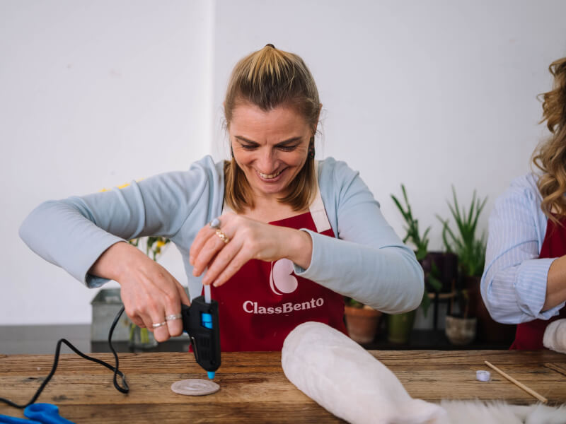 Woman wearing apron smiling whilst using glue gun at a craft class.