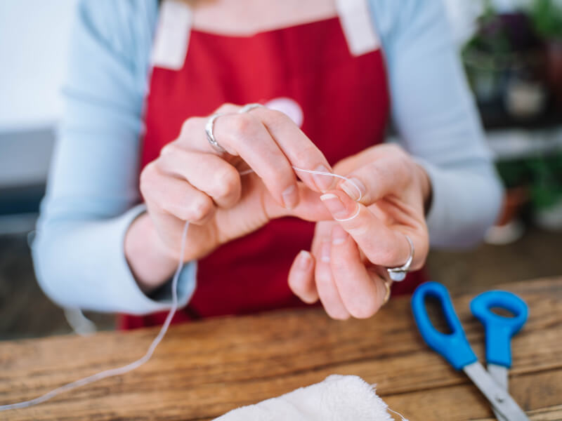 Hands holding a white thread next to a pair of blue scissors. 