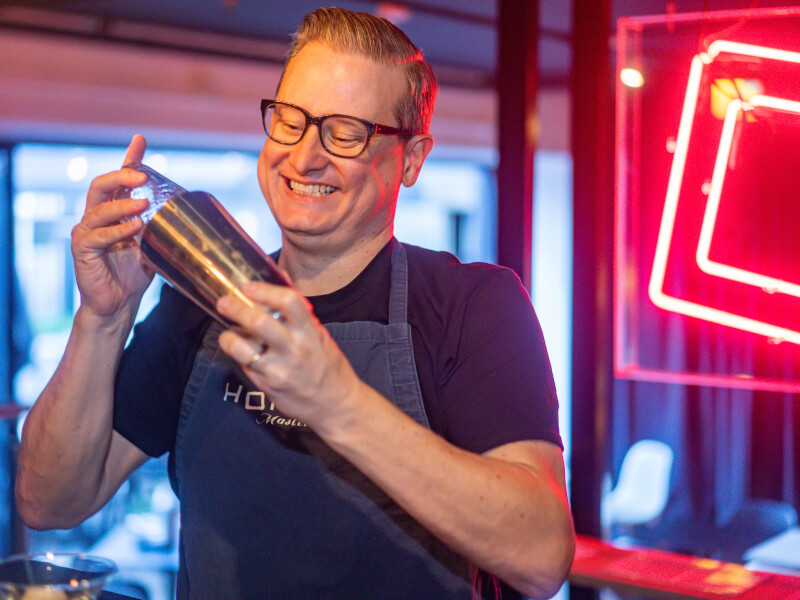 A smiling man is standing behind a bar, using a cocktail shaker at a cocktail making class