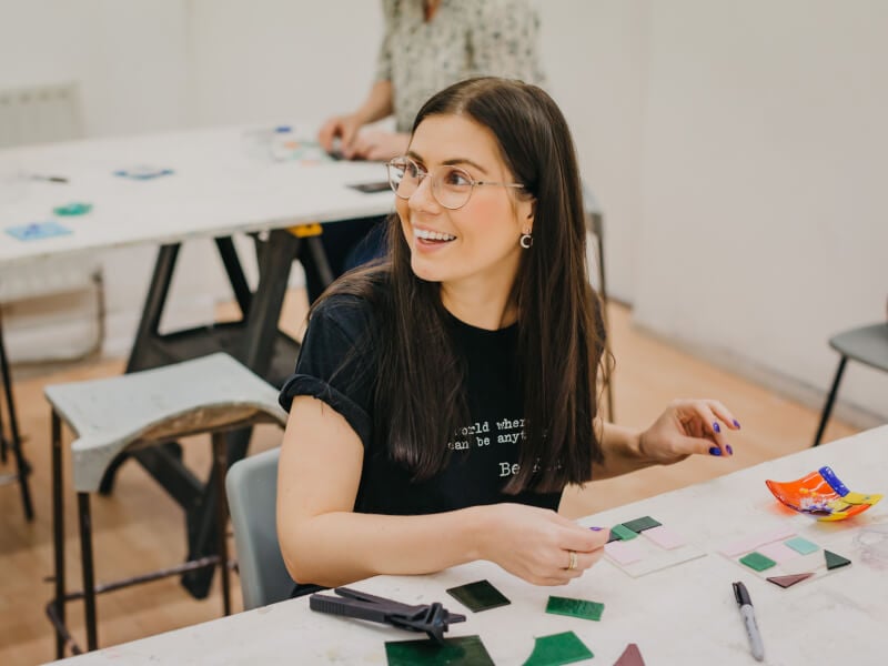 A woman in silver glasses smiling whilst placing mosaic pieces in pink and green onto a glass tile