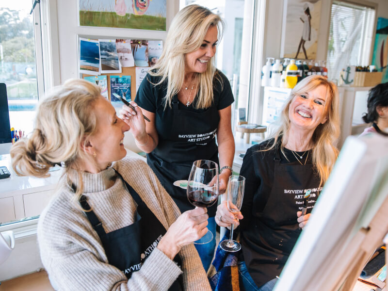 A group of women are sitting at easels laughing with drinks at a wine painting class