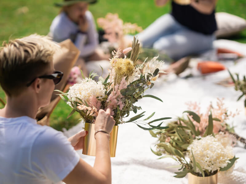 A woman is sitting outside at a table, creating a dried floral arrangement