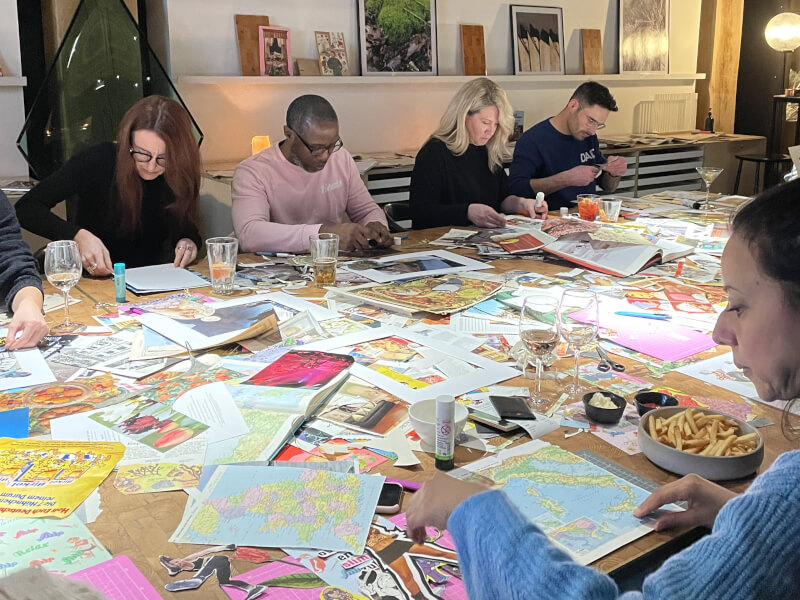 A group of people sit at a large wooden table, making artwork using collage techniques