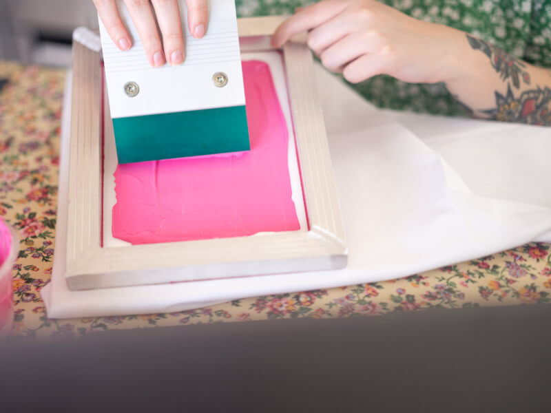 A person using a small printing screen to print on cloth with pink ink