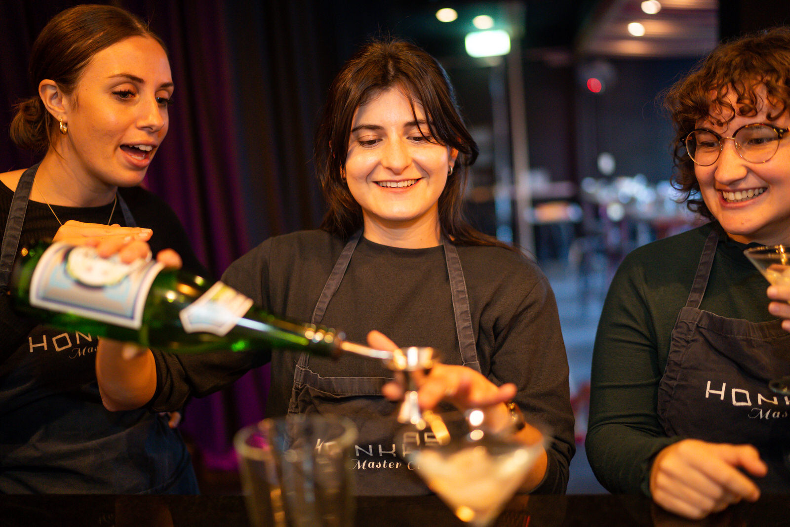 Three people in aprons smiling and talking as they measure out cocktail ingredients