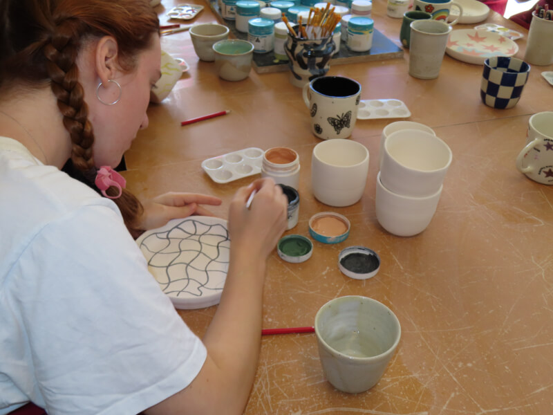 Over the shoulder view of a person painting a bowl with a variety of coloured paints
