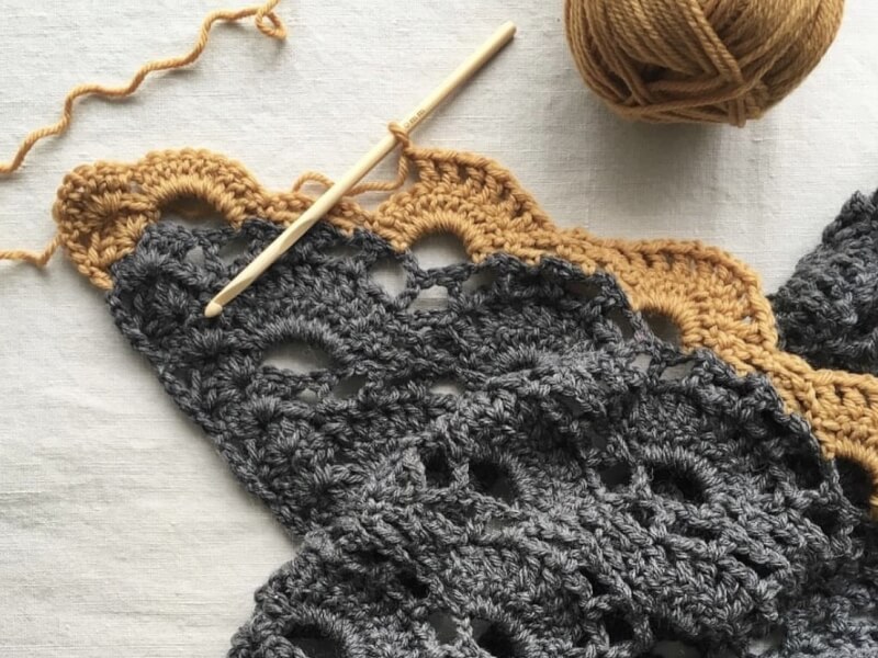 A grey and yellow crochet shawl with a crochet needle