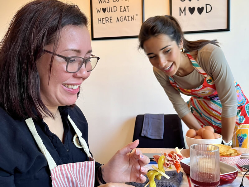 Two women are standing at a wooden table, pushing meat and cheese onto a wooden skewer