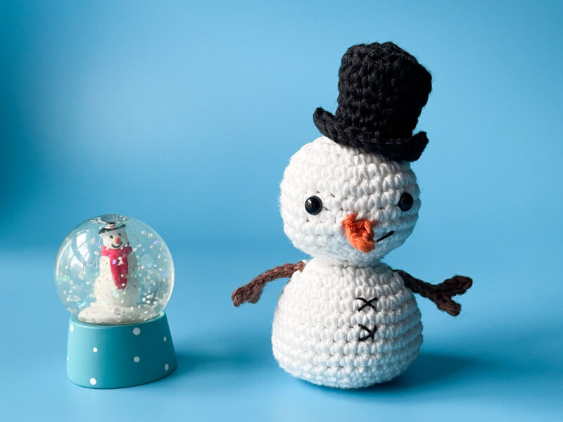 A crocheted snowman sitting on a blue table with a snowglobe next to it
