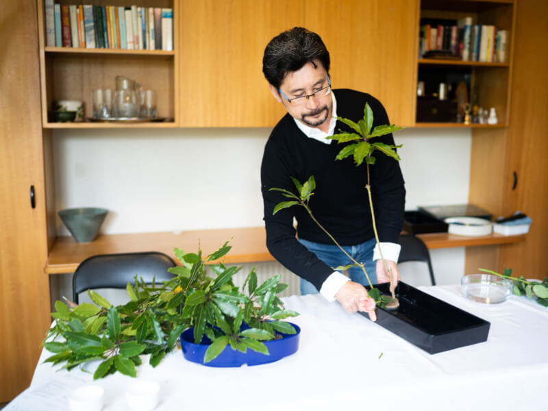 A man in a white shirt and black sweater trimming an ikebana arrangement with shears