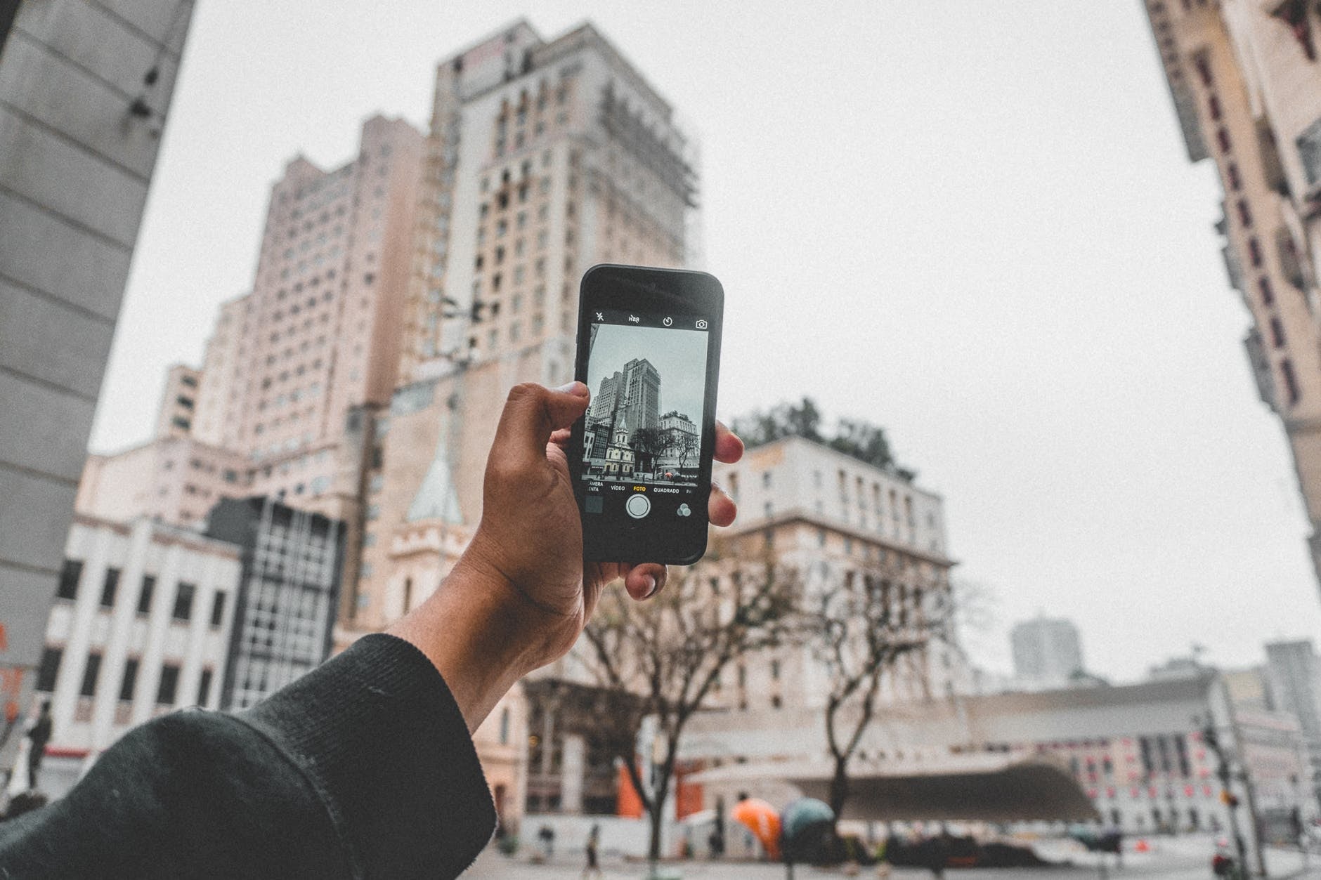 A hand holding a smartphone, taking a picture of tall city buildings