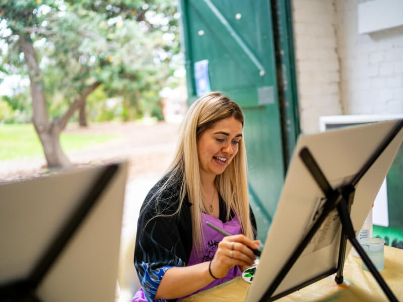 A smiling woman sitting at a paint canvas