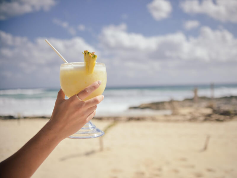 A hand holds up a pina colada cocktail with blue sky and a sandy beach in the background