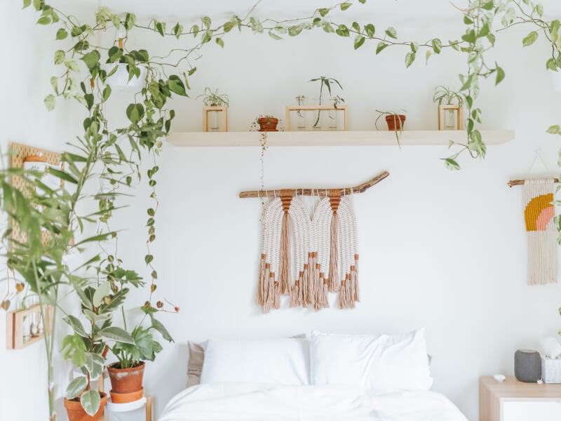 a white bedroom is filled with a bed, macrame wallhanging and lots of plants