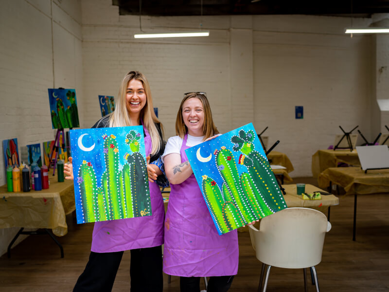 two women holding canvases in a painting class