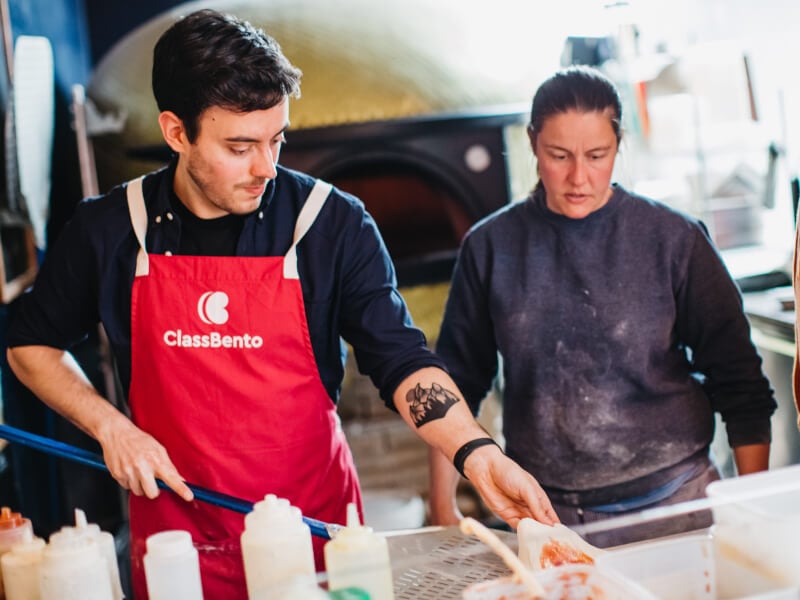  A man in a ClassBento apron moves a pizza base made of fresh dough onto a work surface with a woman assisting beside him