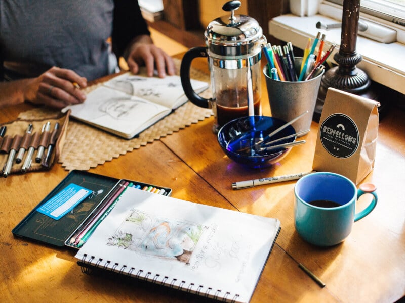 Person sketching in book on a table with coffee pot and paintbrushes
