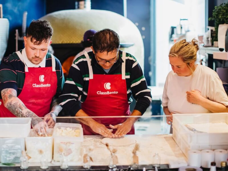 Two men in aprons stand side by side while a woman teaches them how to make pizza dough