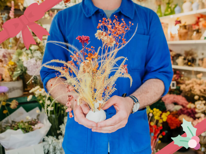 A man holds a dried flower arrangement in bright colours of red, orange and beige