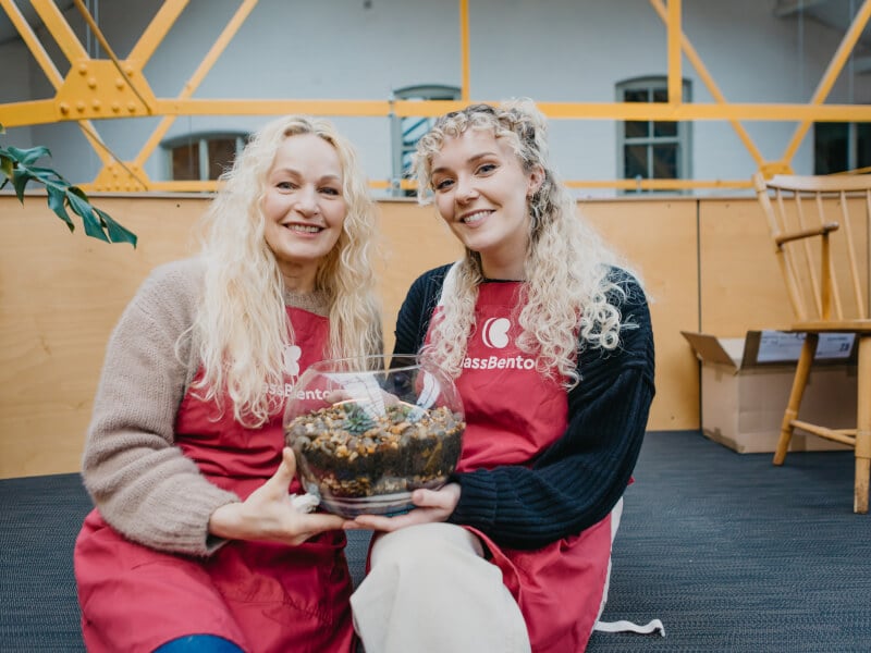 Two women sit wearing red aprons holding a large glass terrarium between them