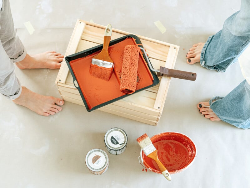 Two people stand next to a paint roller and tray willed with orange paint
