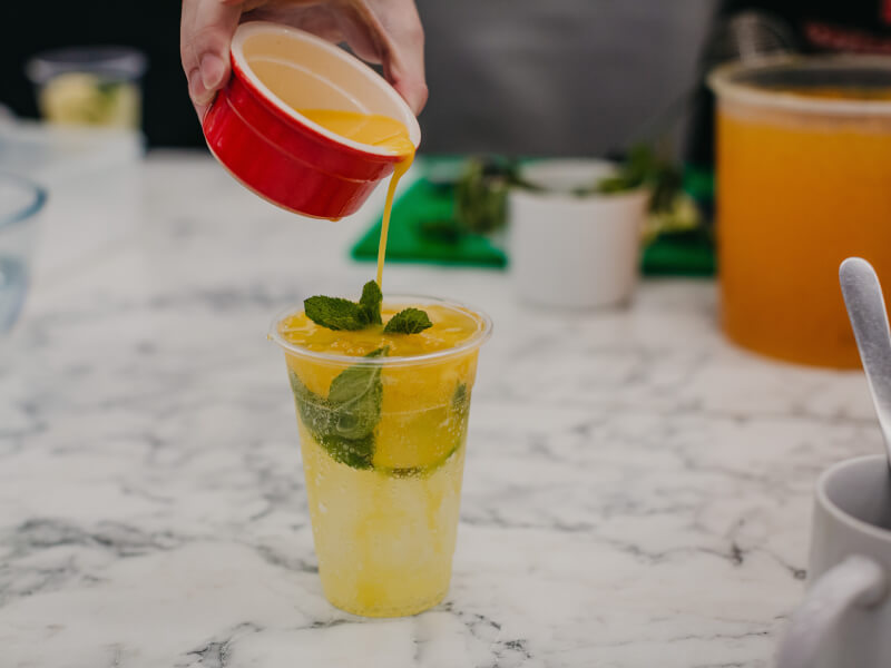 A hand pours an orange syrup into a cup of ginger jasmine mocktail with fresh lime from a red ramakin
