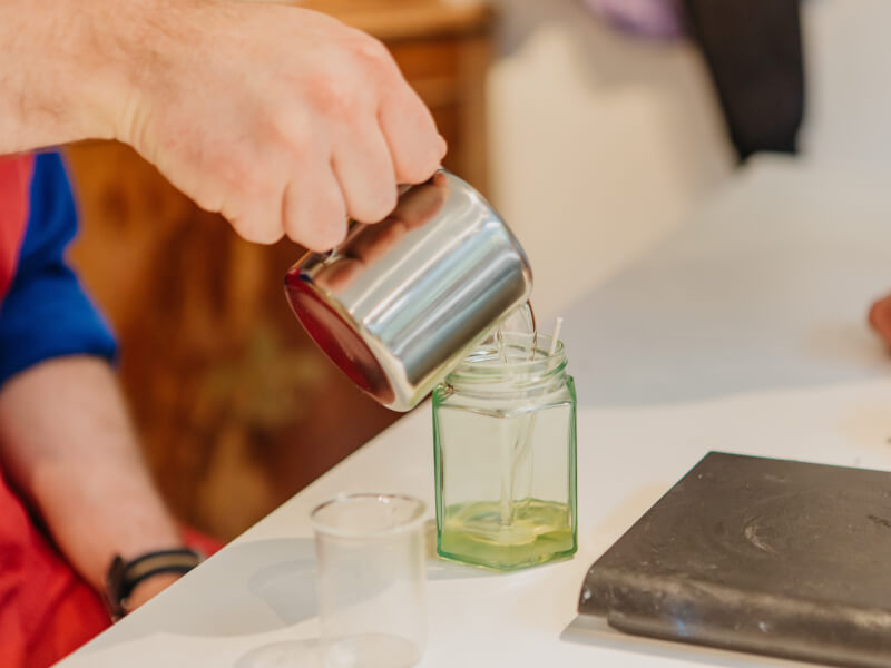 A man pours soy wax into a glass candle jar from a small metal jug