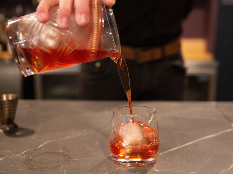 A hand pours an old fashioned cocktail into a small glass tumbler through a cocktail strainer