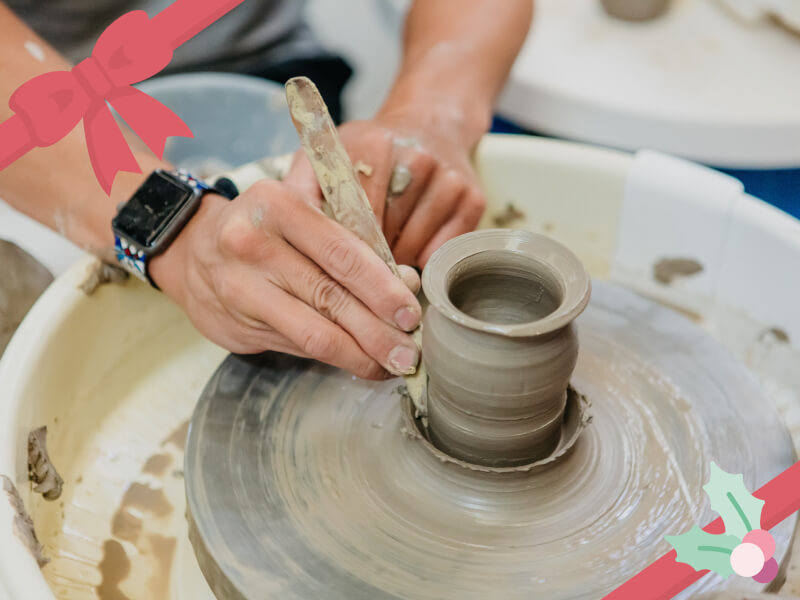 A person's hand holds a small metal tool to an almost complete vase on a spinning pottery wheel