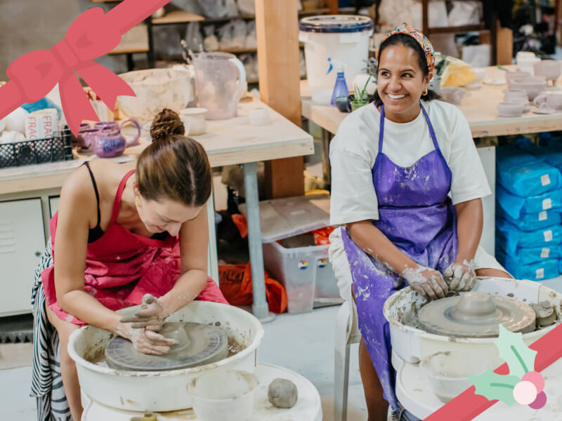 Two women in aprons smile and laugh as they work on pottery wheels