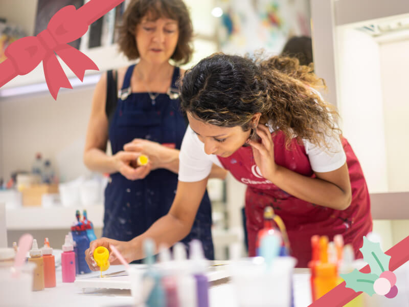 A woman in a red apron leans over a craft table to pour some paint onto a canvas with a tutor looking on behind