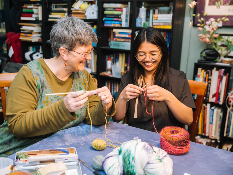 Two women are sitting at a table at a crochet class, working with coloured yarn and wooden crochet hooks
