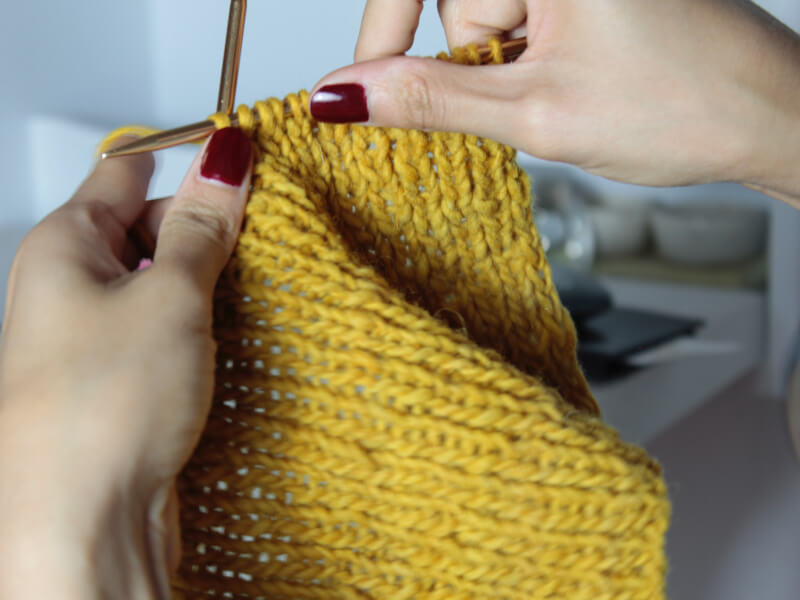 A close up of a person knitting with gold yarn and wooden knitting needles