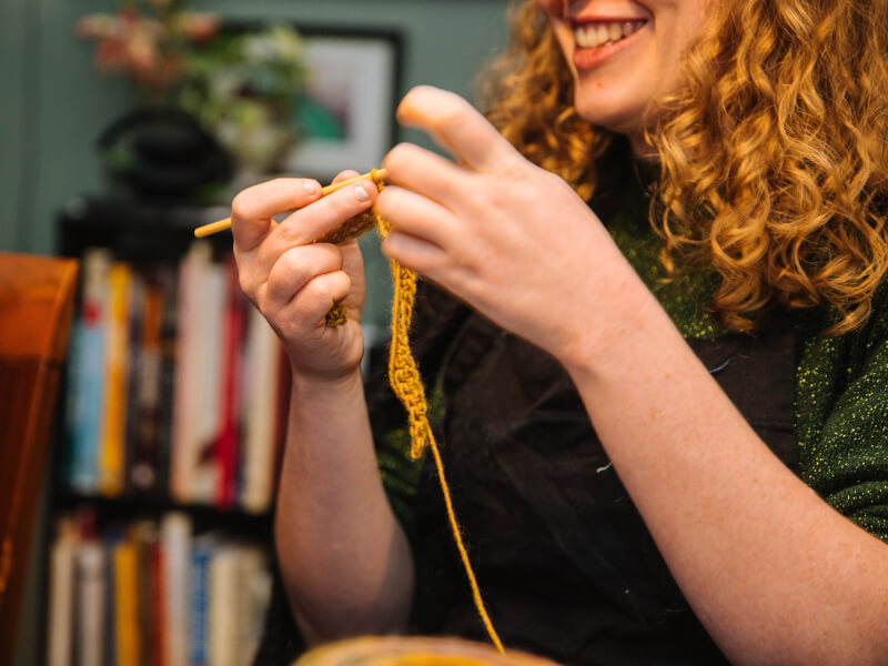 A woman is smiling and learning to crochet with a wooden crochet hook and gold coloured yarn