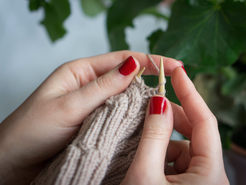 A close up of a persons hands knitting in beige yarn with wooden knitting needles