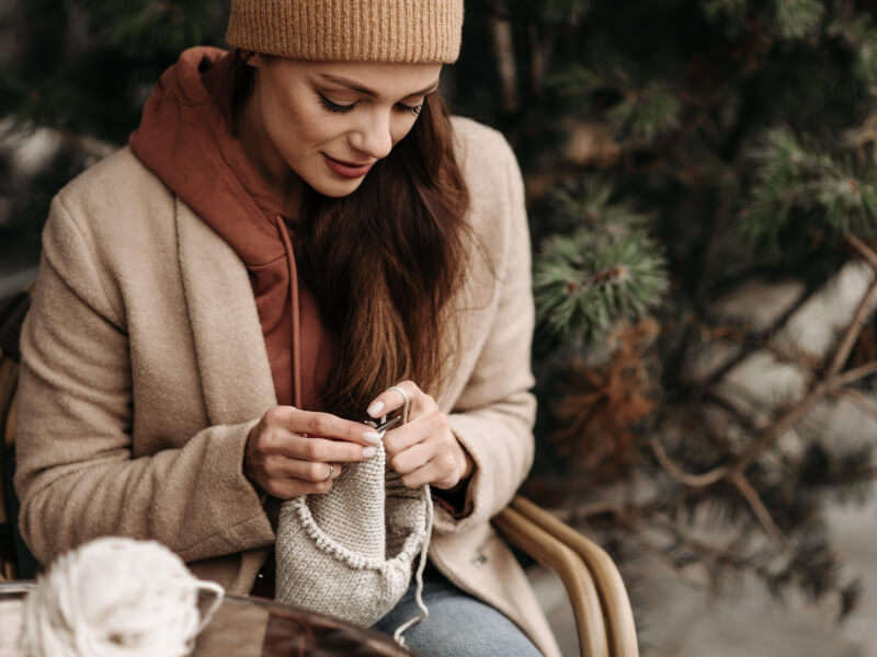 A woman sits at a table outside a cafe, knitting a project in the round in cream yarn