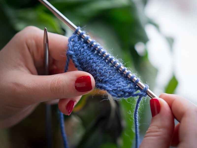A close up of a person with red fingernails working on a knitting project in blue yarn