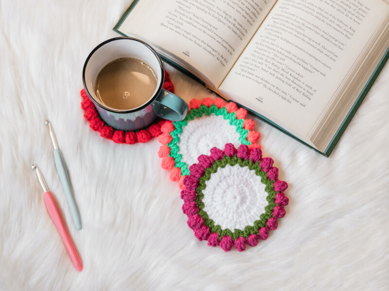 A top down shot of crocheted coasters in white yarn, edged with green and pink decorative crochet stitches
