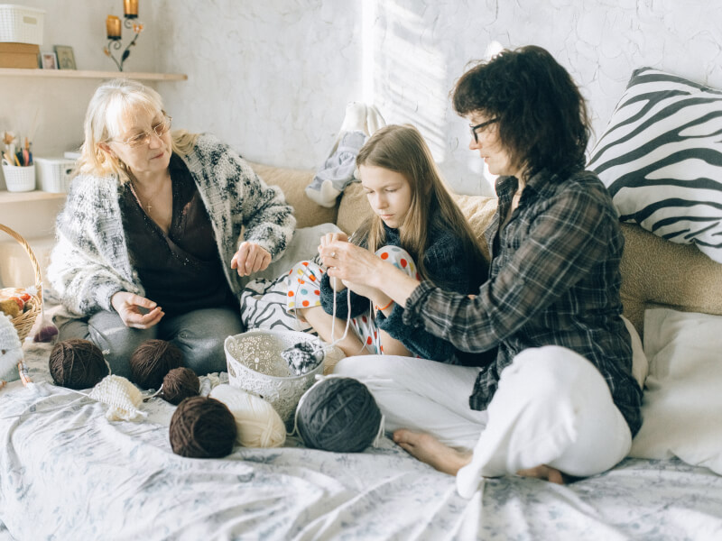Two women are teaching a young girl to knit with grey and white yarn