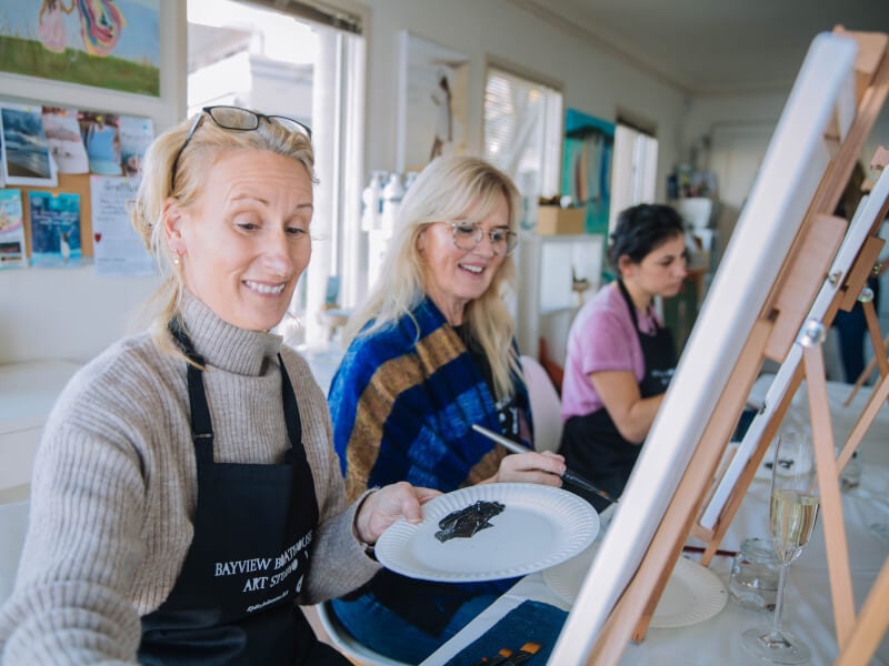 Three women sitting in front of paint canvases smiling.