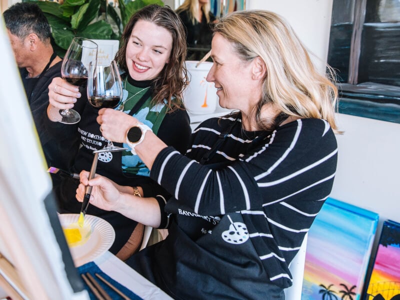 Two women sitting at easels are clinking glasses at a paint and sip class